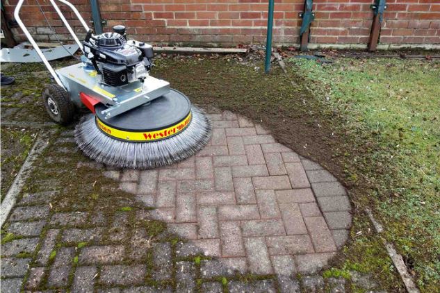 Westermann power sweeper cleaning a red brick patio, brushing away dirt and grass, against a brick wall and lawn.